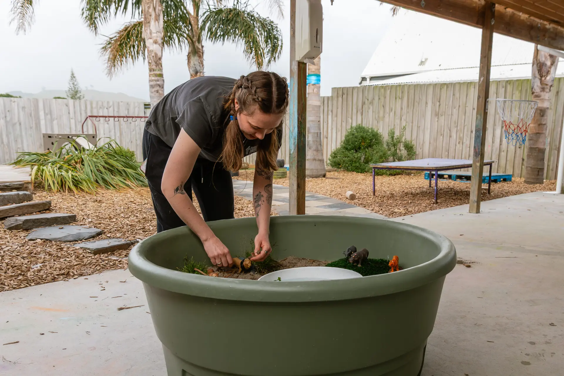 Teacher leaning over a Fern-green All-in-One PlayTray set up with natural materials at an outdoor early-learning centre